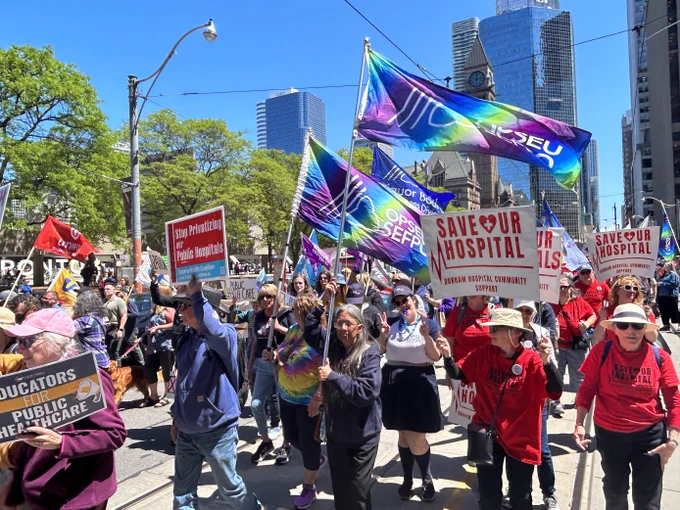 A photo of a large crowd of marching trade unionists, holding oPSEU/SEFPO flags, "Save Our Hospital" placards, "Educators for Public Healthcare" placards, and other union flags. The city buildings of Toronto, including the large "TORONTO" sign outside City Hall, are visible in the background.