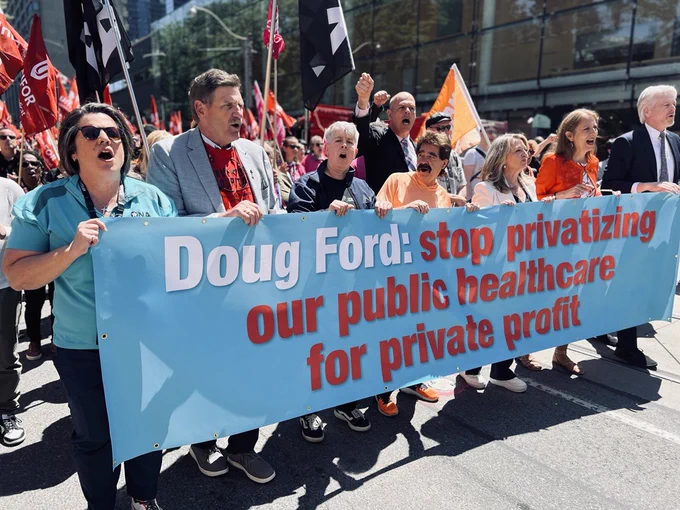 A group photo of trade unionists holding a blue banner with white and red writing which reads, "Doug Ford: stop privatizing our public healthcare for private profit!" OPSEU/SEFPO President JP Hornick holds the banner near the middle of the front line. Visible behind is a large crowd marching with union flags, including from Ontario Nurses Association and UNIFOR.