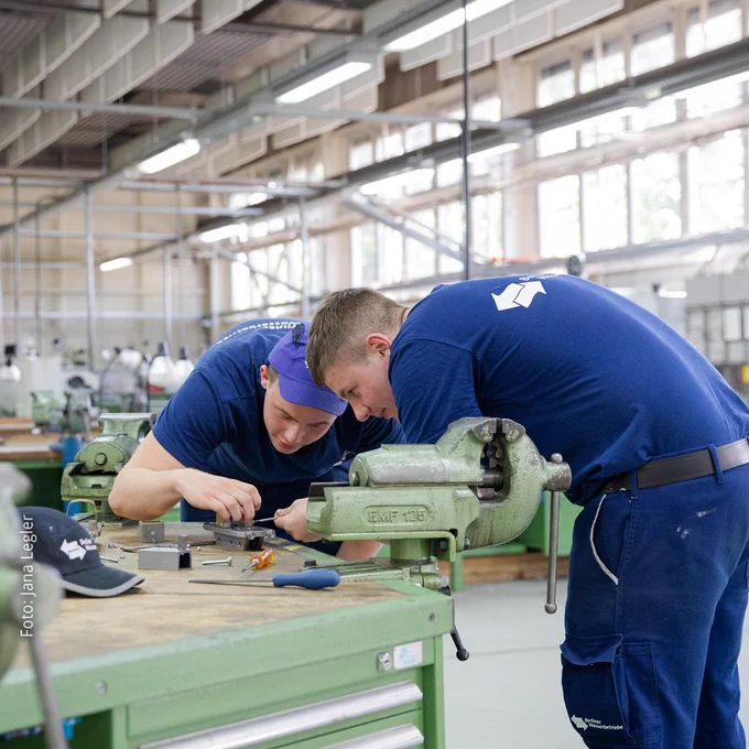 Foto von zwei Auszubildenden der Berliner Wasserbetriebe, die an einer Werkbank in der Werkstatt an einem Metallgegenstand arbeiten.
