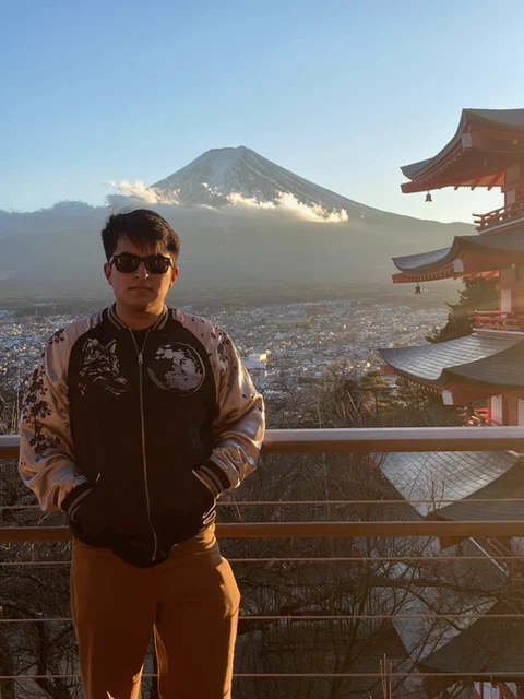 An individual stands in the foreground with a scenic view of Mount Fuji and a red pagoda in the background. This person is wearing a dark jacket with a detailed pattern, sunglasses, and mustard-colored pants. The atmosphere is clear, allowing for a vibrant depiction of the landscape.