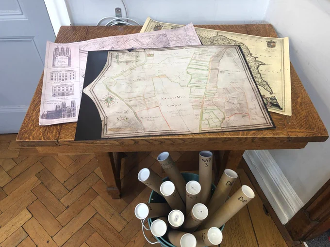 A photograph of a table in the library shop with several maps spread out on it. On top is 1624 Dringhouses map. Under the table is a bucket full of rolled up maps.