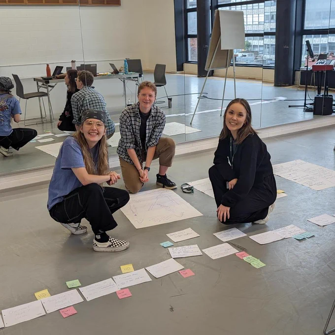 Three individuals in a spacious room with large windows, engaging in a creative planning session on the floor. They are surrounded by various colorful papers and notes spread out. All are smiling, suggesting a positive and collaborative atmosphere.