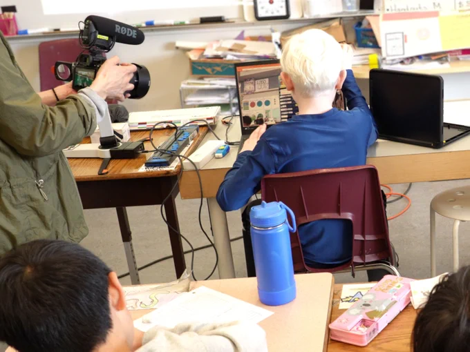 View from behind a child sitting at a desk in a classroom with magnified video screen in front of them. A person on their left holds a video camera filming. Other children at desks are also in view.