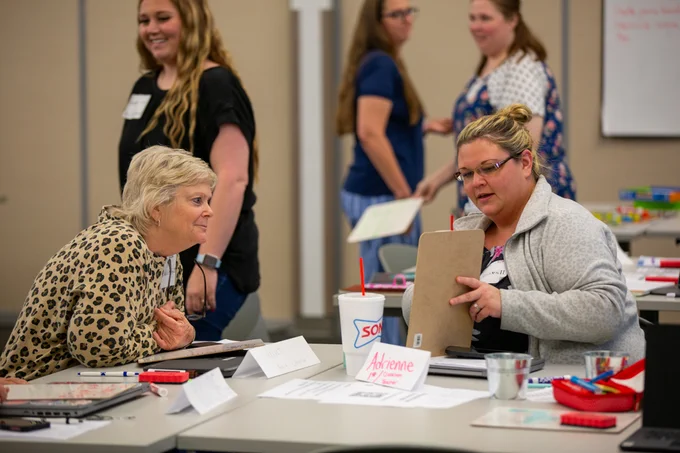 Two educators share their hand-drawn instructions for creating a PB&J sandwhich. One leans over to table, listening intently, while the the other shares her ideas.
