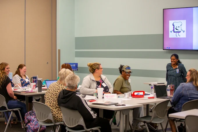 Two educators share their hand-drawn instructions for creating a PB&J sandwhich. One leans over to table, listening intently, while the the other shares her ideas.