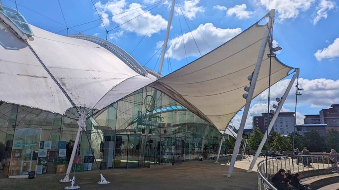 Alternative text description: Exterior view of the Dynamic Earth science center in Edinburgh, featuring its distinctive white tensile canopy structures against a backdrop of a blue sky with scattered clouds.