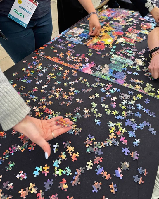 People are collaboratively assembling a large, colorful jigsaw puzzle on a table.
