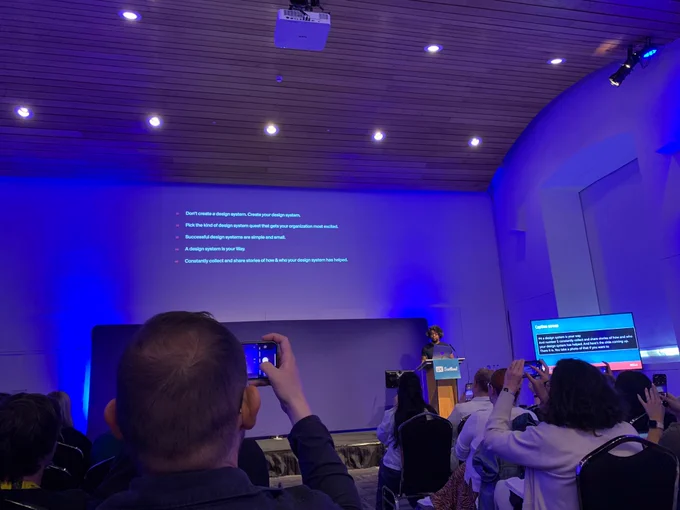 Audience members taking photos of Dan Mall on stage at UX Scotland with visible presentation screens 