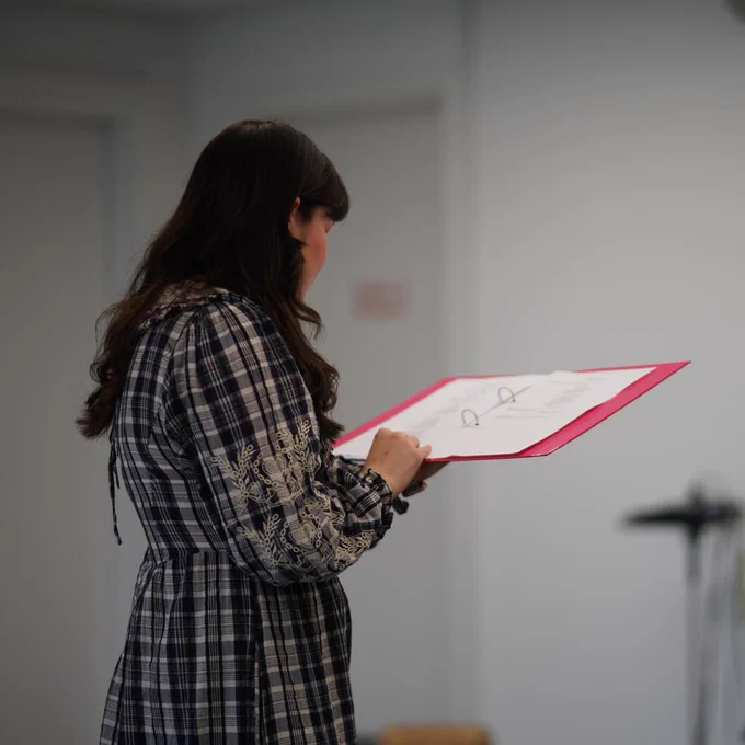 Hannah stands in a room fading away from the camera, holding a script.