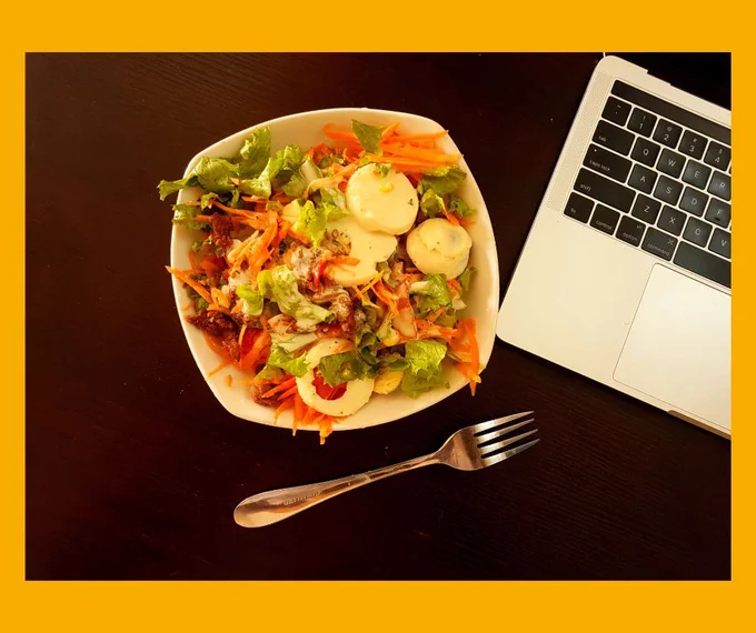 A salad in a white bowl next to the laptop with a silver fork to one side