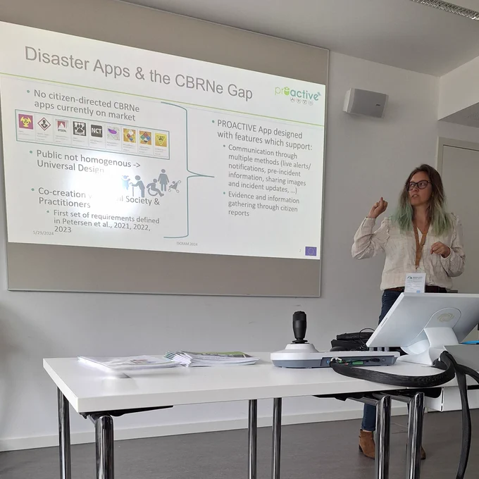 White women with blond and blue hair, wearing glasses and casual business attire, with the ISCRAM conference landyard on, is gesturing towards the powerpoint slide project. The slide reads "Disaster Apps & the CBRNe Gap"
