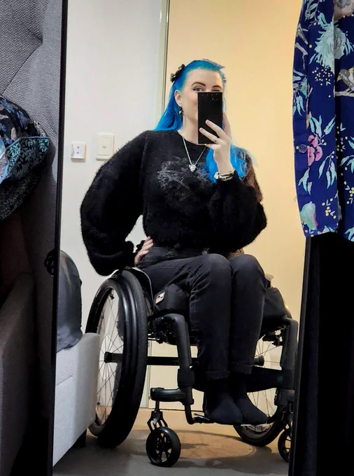 A self-taken photograph of Dragancaor, a white nonbinary femme person with green eyes & long blue hair, sitting in their black, custom wheelchair in front of a full length mirror. The wheels have a slight camber & there is no visible backrest (as it is low & stops at their waist). They are wearing black jeans & a black fluffy cropped Killstar jumper.