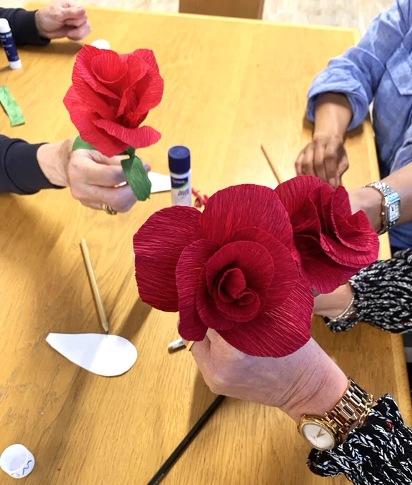 Red paper crafted roses held up towards the camera by three attendees to the Traditional Paper Flower craft event at Yiewsley library. The attendees are sat around a table upon which there are other craft materials including a glue stick, pencils and paper. 