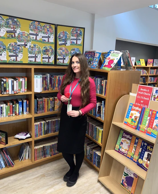 Roksana, staff member at Yiewsley Library stood within the library facing the camera holding a paper crafted rose. Roksana is wearing a red top and black skirt with her long hair down. Behind her are bookcases and a tree background display. 