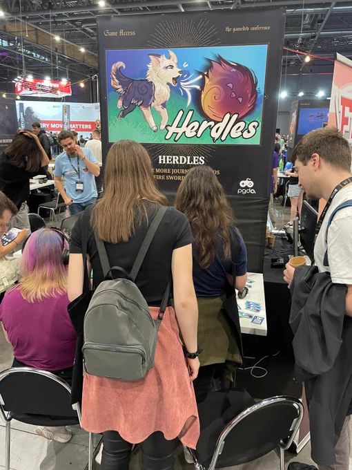 A group of people standing around a PC setup where two people are playing our game Herdles. 