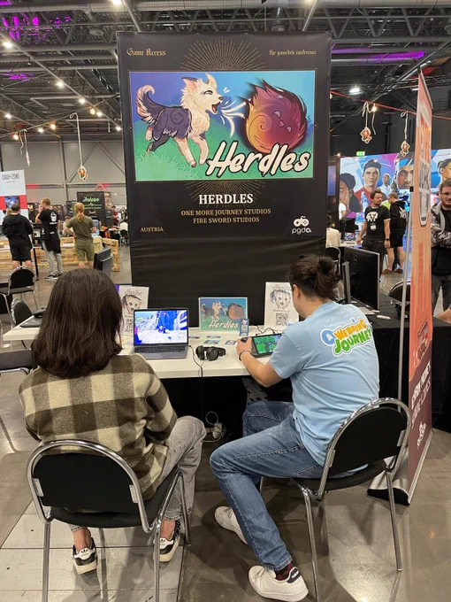 Two people sitting at a table playing our game Herdles. One person playing with a controller on a MacBook and the other one holding a steam deck.
Above the table is a banner with the key artwork of the game. 