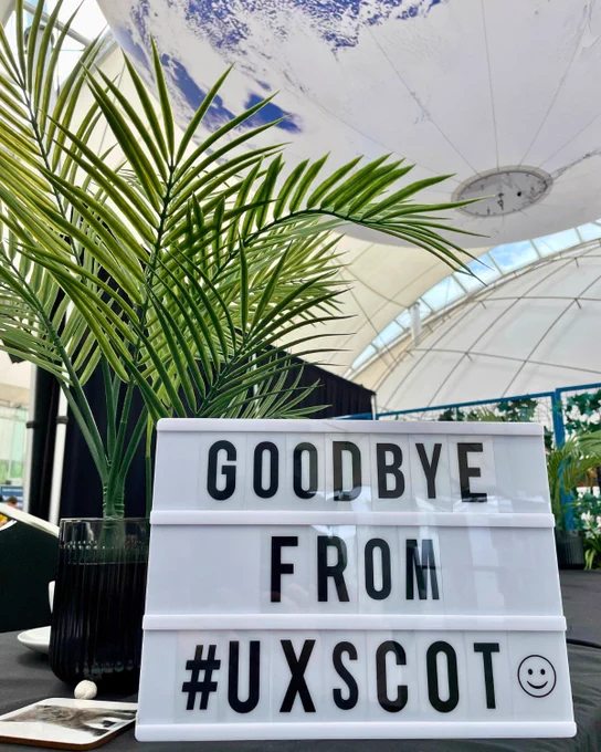 Lightbox sign with the text "GOODBYE FROM #UXSCOT :)" in front of a large globe and a potted palm plant, displayed in a venue with a translucent roof.
