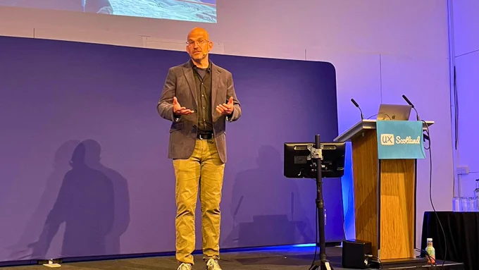 Jeff Gothelf presents onstage at a conference, standing at a podium with the sign "UX Scotland" in front of a large screen displaying a digital image.