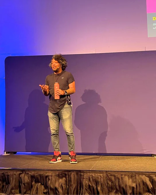 Dan Mall standing on stage, dressed casually in a t-shirt, jeans, and sneakers, presenting in front of a large purple backdrop and a brightly colored display with text.