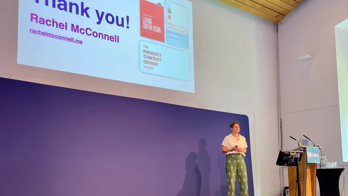 Rach McConnell standing on stage at a conference beside a large screen displaying a presentation slide that reads, "Thank you! "