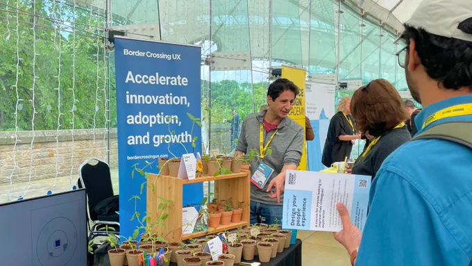 A trade show booth labeled "Border Crossing UX" with a banner that reads "Accelerate innovation, adoption, and growth" in a tented area. The booth features multiple potted plants on display and a staff member engaged in conversation with participant.