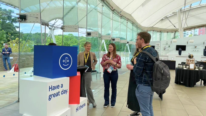 Four individuals conversing at the Tesco Bank booth with welcoming signs that read "Have a great day" and "Hi there," set up under a large, white canopy. The background shows lush green trees and a cloudy sky.