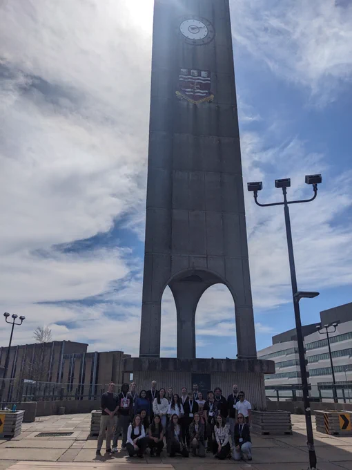 Twenty-three people are standing/kneeling in front of the Memorial University clock tower. They are smiling at the camera. The sky is lightly cloudy.