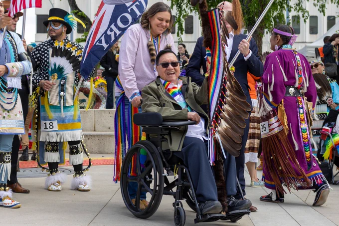 A powwow participant in a rainbow ribbon shirt holds a staff from a wheel chair in Churchill Square. A helper looks over the participant from behind the chair. Several others surrounding the pair are dressed in regalia.