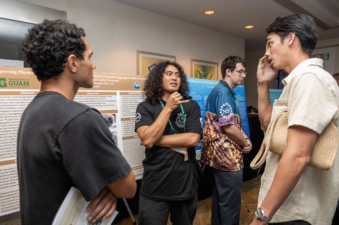 Three students stand in a circle having a conversation in front of one of the student's research poster. 