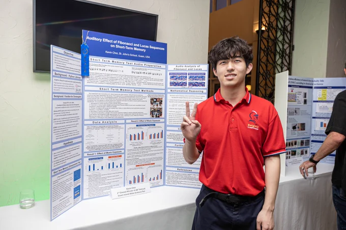 A student wearing a red polo shirt holds up a peace sign in front of his research trifold poster. 