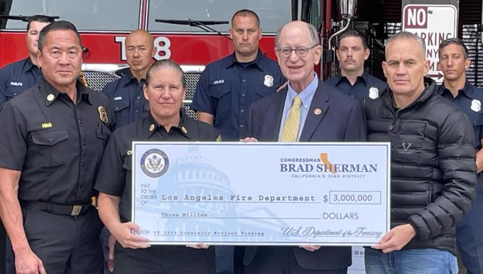 Standing in front of the rear apparatus bay doors at LAFD Station 88 in Sherman Oaks, with the  cab of an aerial ladder truck prominently in the background behind five firefighters in their work uniforms, is (left to right) LAFD Deputy Chief Jason Hing, LAFD Fire Chief Kristin Crowley, U.S. Congressman Brad Sherman, and IAFF General Secretary-Treasurer Frank Lima, the latter three holding a large presentation check payable to the "Los Angeles Fire Department" for the amount  of $3,000,000. 