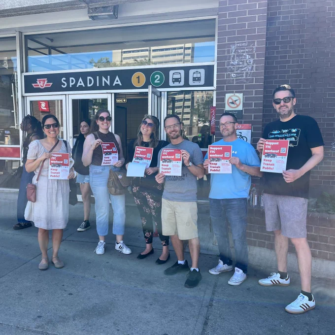 Six people are standing in front of Spadina TTC station. They are all holding signs that say: SUPPORT TTC WORKERS! KEEP TRANSIT PUBLIC! 