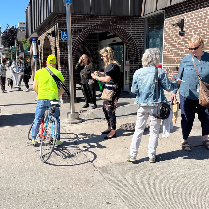 In this photo, many conversations are underway. Two people are handing out leaflets, one person is taking one, another has just signed the petition. In the background, another conversation is clearly underway, with one person holding leaflets, and two others engaged. There is a cute dog on a leash while the other talk. 

