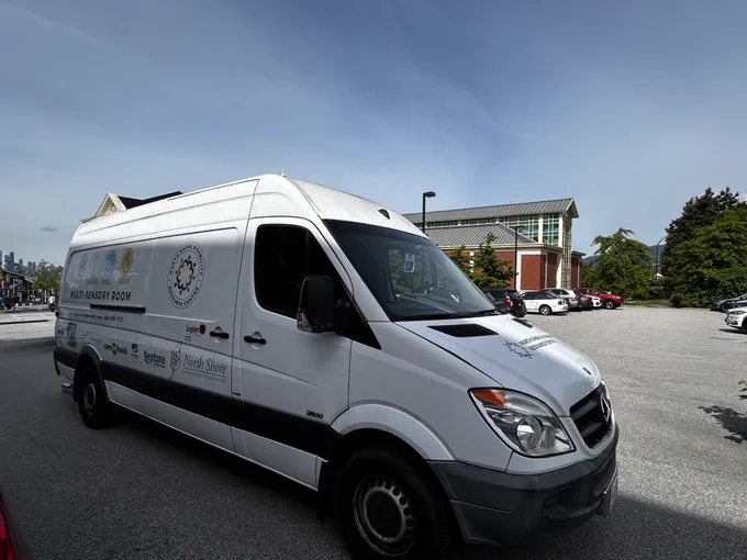 A white van with logo decals across the side is parked at the end of a paved parking lot beside a school. The sky is blue with a layer of light clouds.