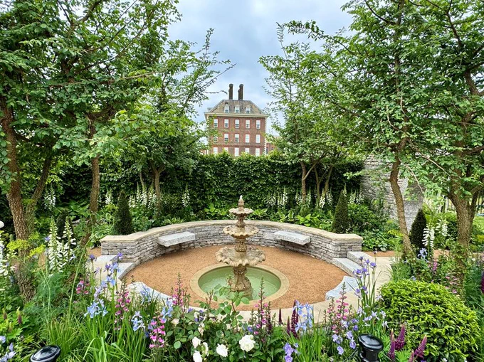 A walled garden with a house in the background and a fountain in the middle of the garden. 