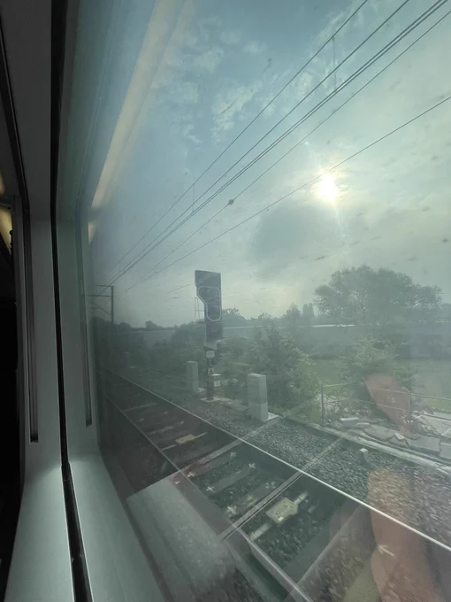 A train window showing a sunny day outside and a passing train track with trees and buildings in the distance