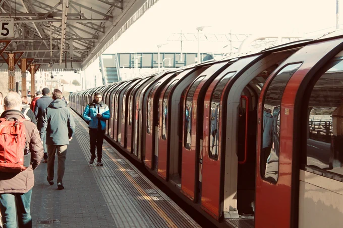 A London tube train sitting at a platform with it's doors open. People are shown walking on the pavement. Tactile marking are prominent on the platform. 