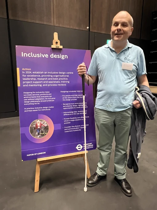 Liam O’Carroll, London SLC coordinator, standing next to a purple board with information about inclusive design. Liam@ks wearing his pale blue sight loss councils t-shirt, holding his long cane, and smiling at the camera.