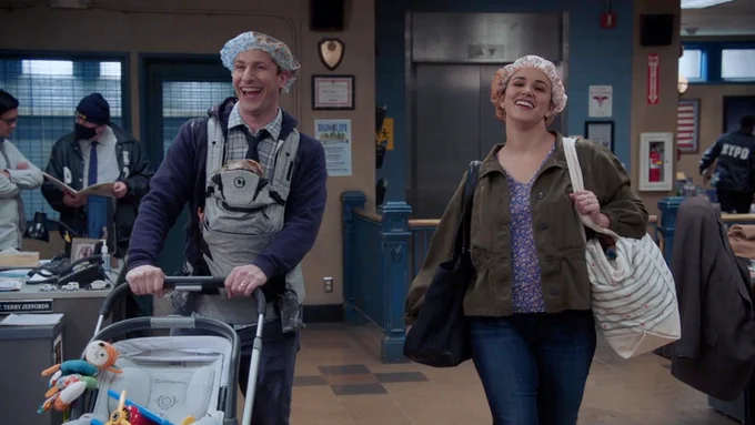 Photo of Andy Samberg as Jake Peralta and Melissa Fumero as Amy Santiago in Brooklyn Nine-Nine. Jake is on the left pushing a stroller, wearing a baby carrier with a shower cap on his head. Amy is on the right holding two bags with a shower cap on her head. They're both smiling.