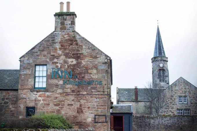 Image of the side of an old brick building with chimney in the foreground with the words Inn at Kingsbarns on the side. A church spire is in the background of the image