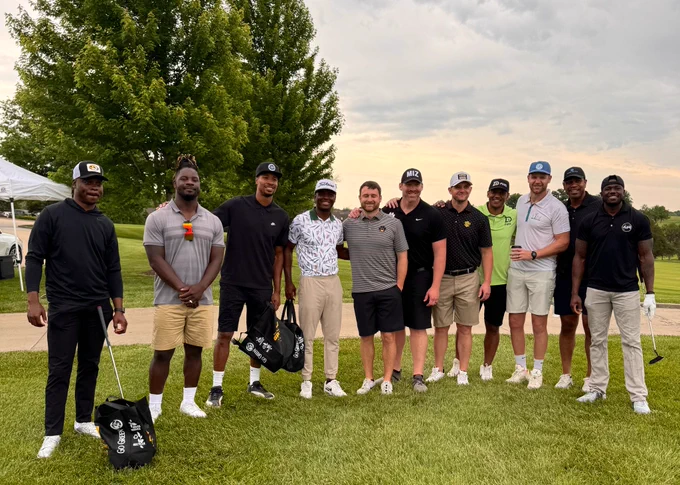 Eleven men stand on green grass in golf attire.