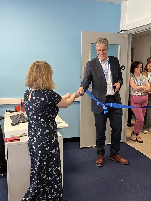 Photo of a woman handing a pair of scissors to a man who is holding a blue ribbon in his other hand. A small crowd of people can be seen standing behind the man in a doorway.