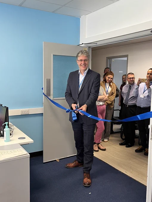 Photo of a man about to cut a blue ribbon. He is holding scissors in his hands in front of the ribbon and smiling towards the camera. A small crowd of people can be seen standing behind him in a doorway.