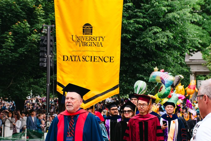  Dean Bourne leads graduating Data Science students to their seats on the lawn, holding up the yellow University of Virginia Data Science banner. 