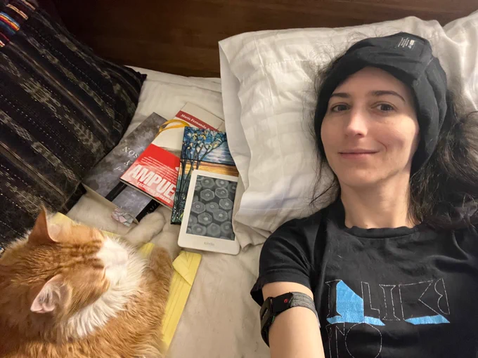 Selfie of a Mexican white woman lying down on a bed with a headache hat on. Near her, an orange tabby resta his paws by multiple books and notebooks.)