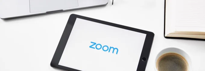 Photo shows a  laptop, tablet displaying the Zoom logo, cup of coffee and open book