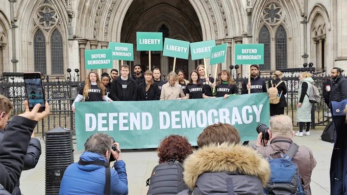 Liberty lawyers and campaigners outside the Royal Courts of Justice holding a banner and placards saying "DEFEND DEMOCRACY" with photographers taking pictures in the foreground