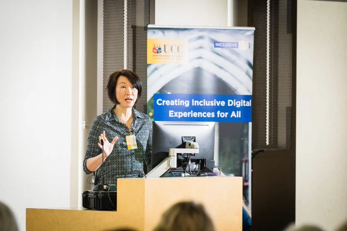 Lilian Joy speaking at the podium with a UCC banner, Creating Inclusive Digital Experience for All.