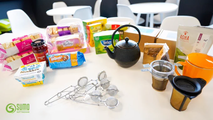 A variety of food items and kitchen tools on a table, including several types of packaged biscuits, jars of jam, a tea kettle, tea bags, measuring spoons, and tea strainers.. The logo "SUMO NOTTINGHAM" is visible on the bottom left side.