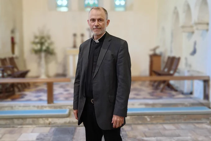 This image shows the Rev David Railton standing in a church. He is dressed in clerical attire, wearing a black clerical shirt with a white collar.  The background features an interior view of a church, with wooden railings, a tiled floor, and an altar.